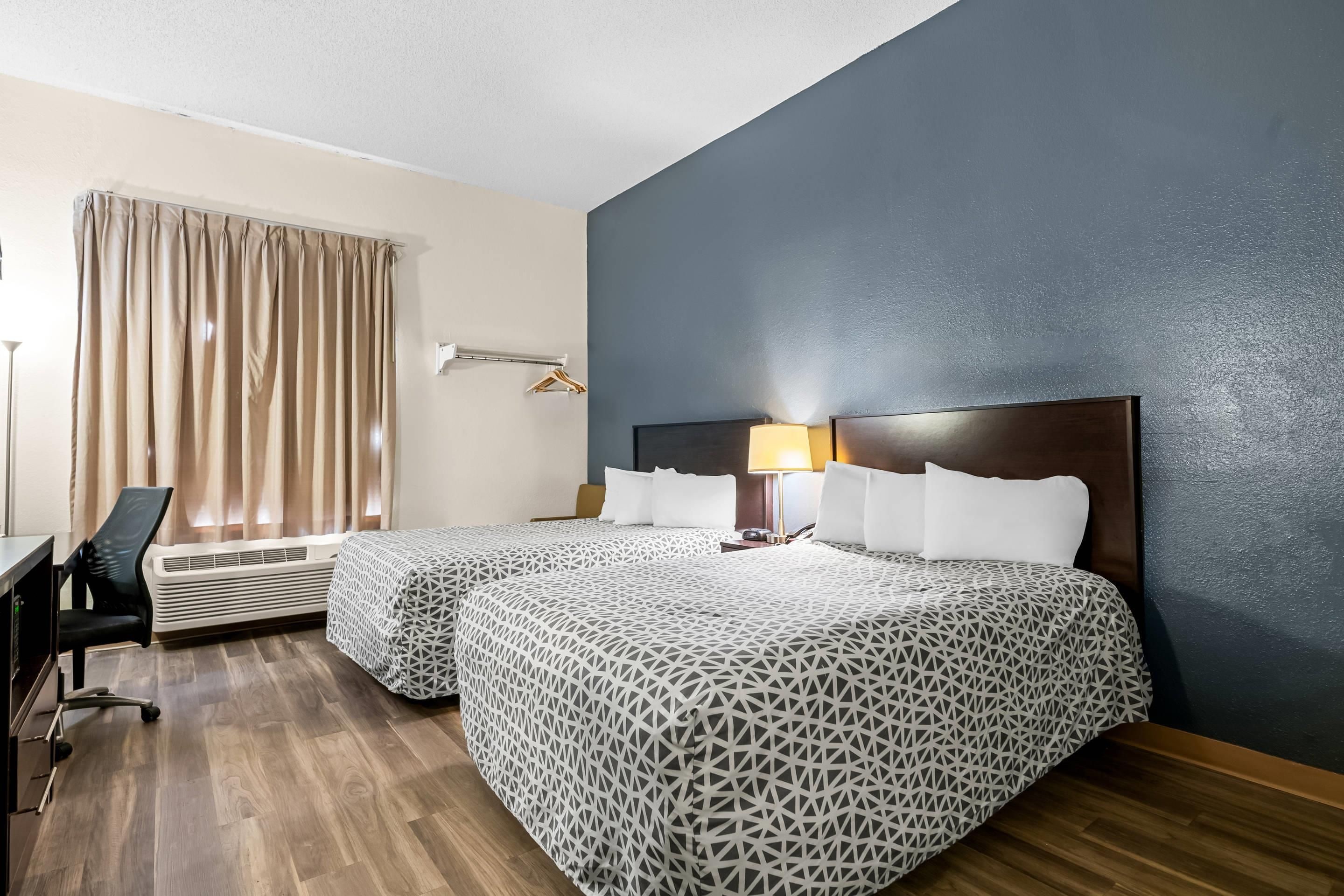 A hotel room with two beds, a desk, a chair, an air conditioning unit, and a window with curtains.