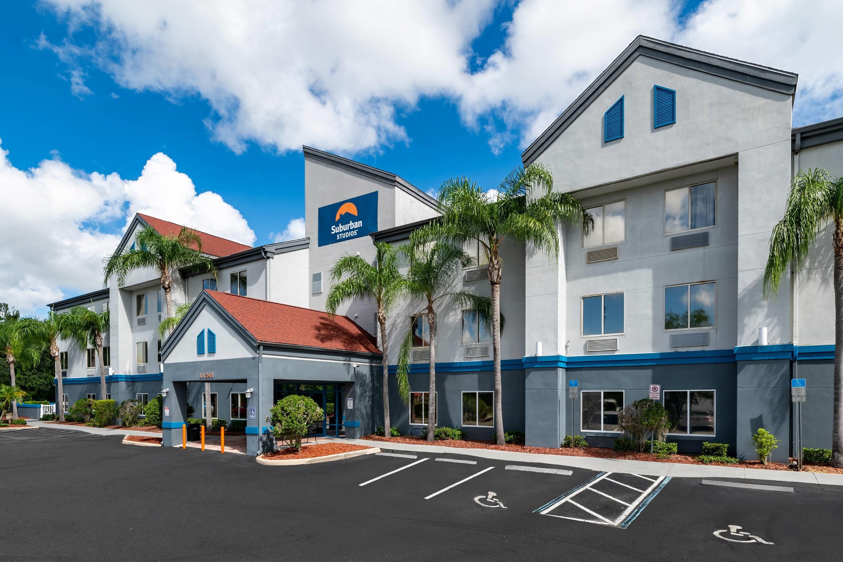 Hotel com design moderno, exibindo uma placa grande com a inscrição 'Suburban Studios', entrada coberta e estacionamento com acessibilidade para pessoas com deficiência.
