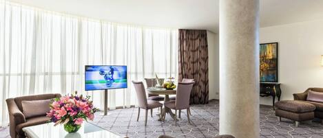 Habitación de hotel con sala de estar que incluye un sofá, una mesa de centro, un televisor, una mesa de comedor con sillas y un ventanal con cortinas translúcidas.