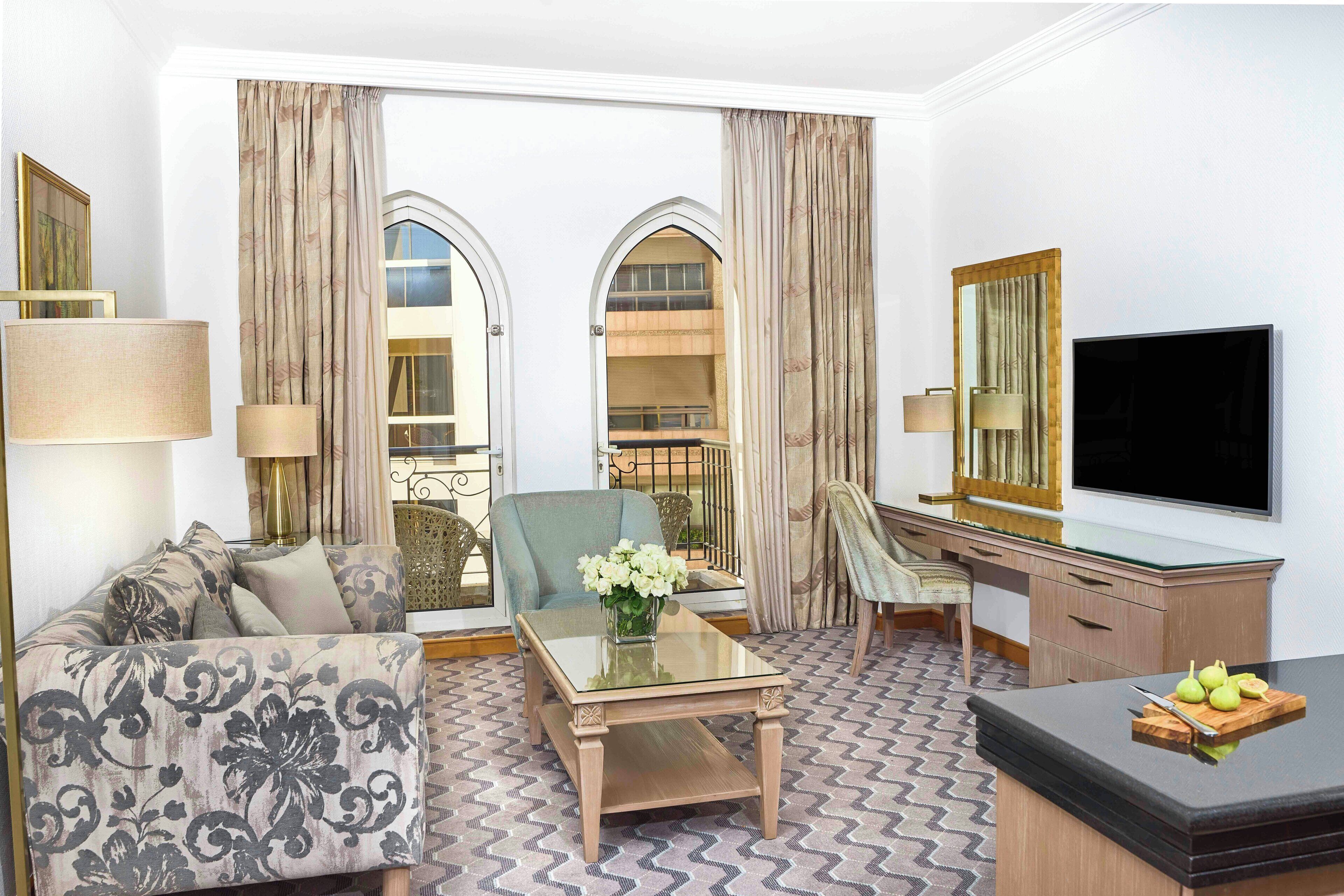 A hotel room with a sofa, armchair, glass coffee table, desk, and a flat-screen TV.