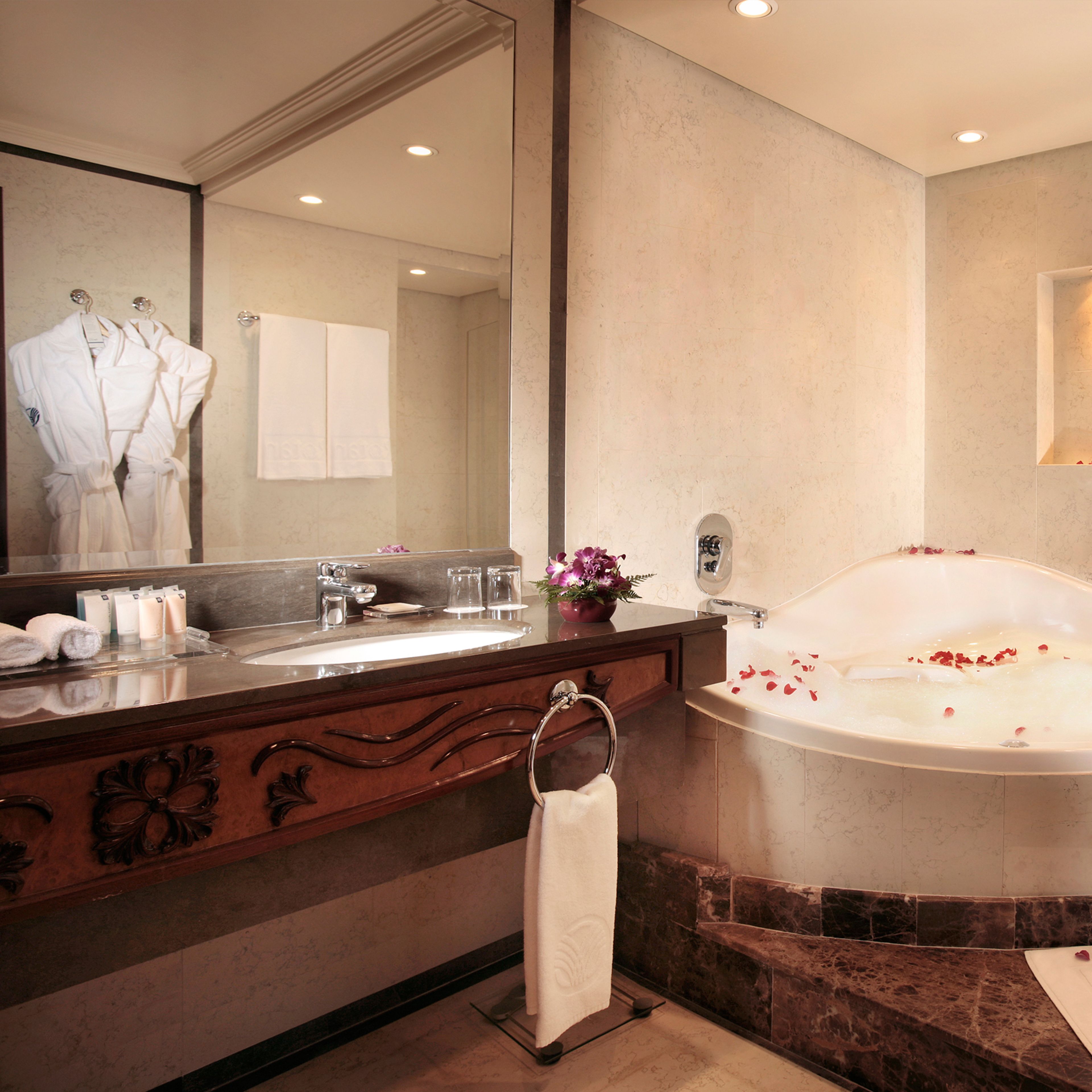 A bathroom with a large mirror, a bathtub with rose petals, a sink with toiletries, and a towel rack.