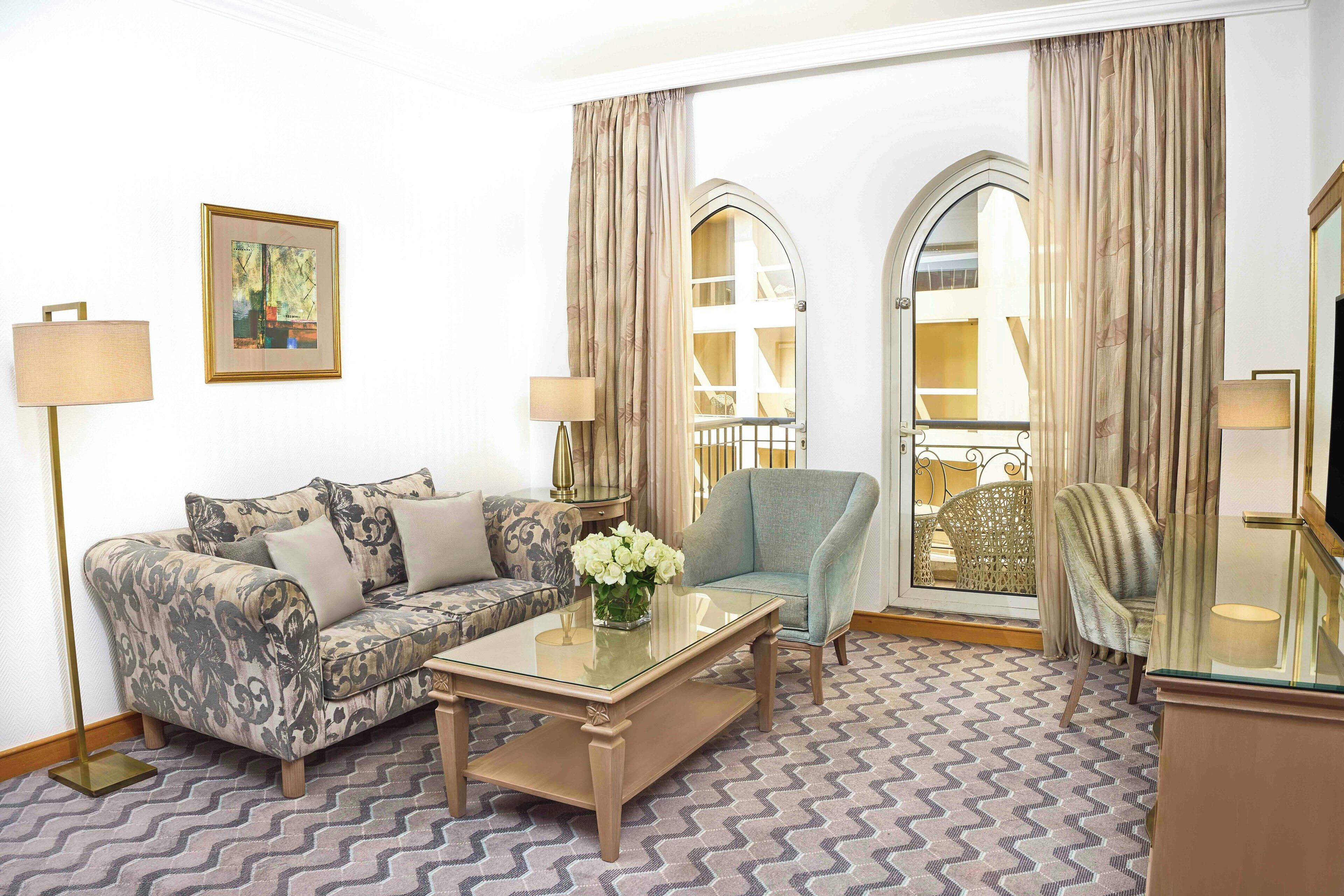 A living room with a patterned sofa, a glass coffee table, a blue armchair, and a decorative vase with flowers.