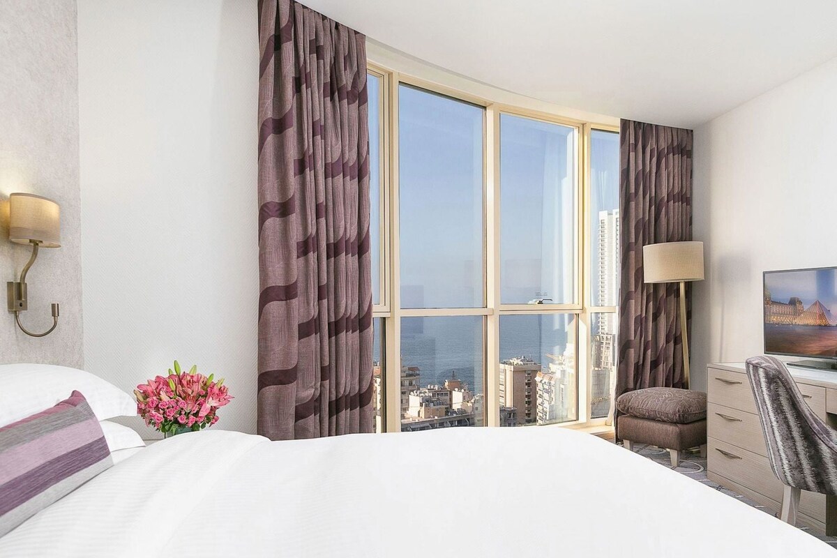 A hotel room with a large bed, a desk with a chair, a television, and a view of the cityscape through floor-to-ceiling windows.