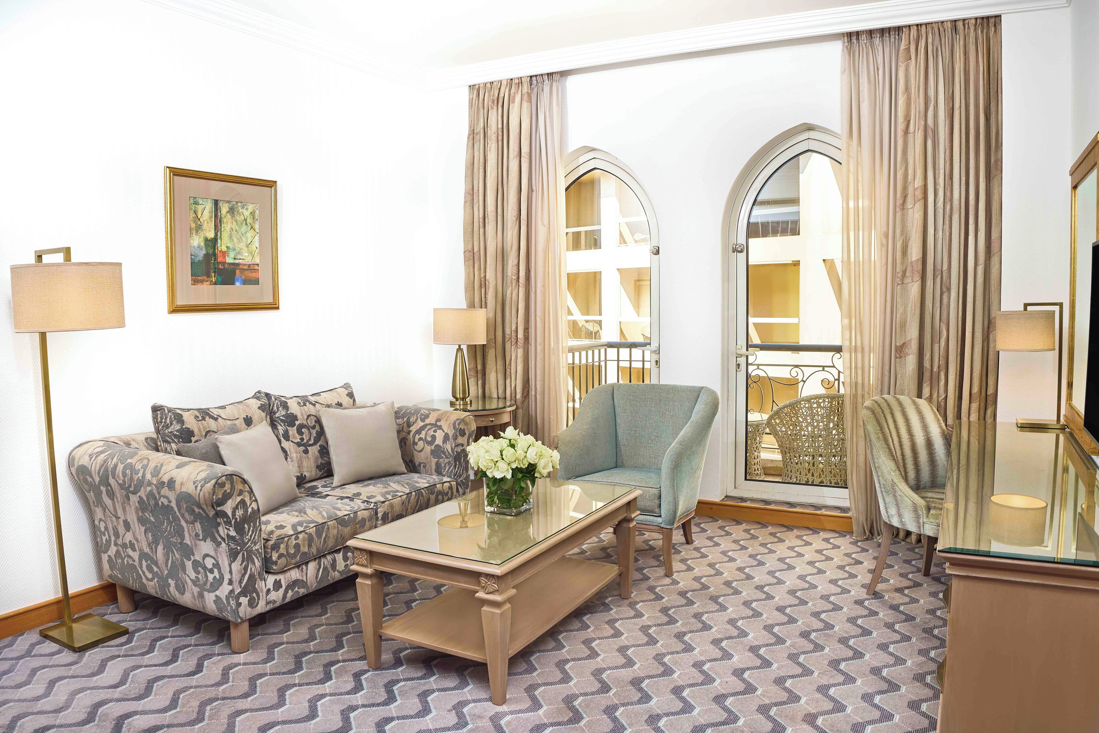 A living room with a patterned sofa, a glass coffee table, a blue armchair, and a decorative vase with flowers.