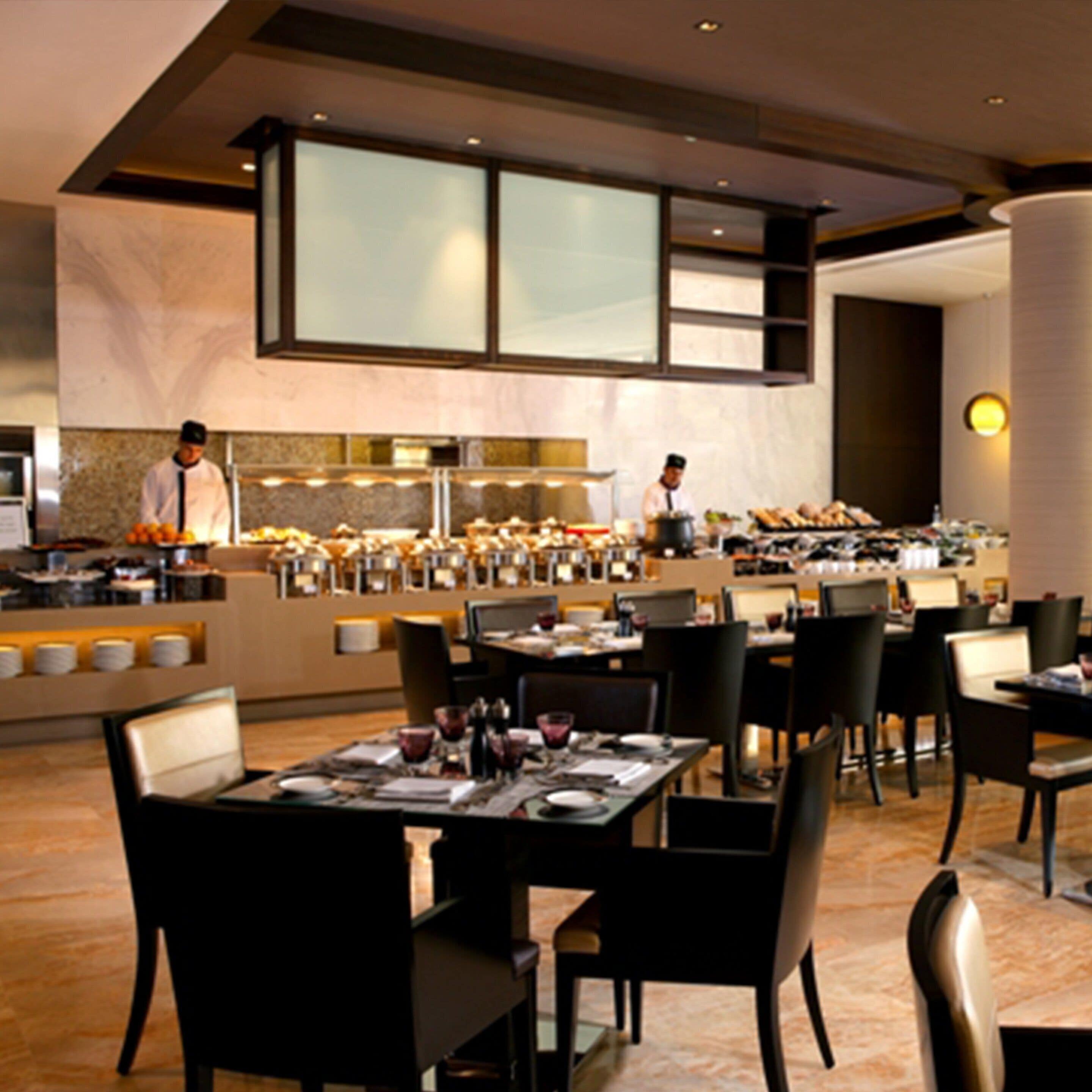 Un comedor moderno con varias mesas y sillas, un mostrador de buffet y una zona de cocina con chefs.