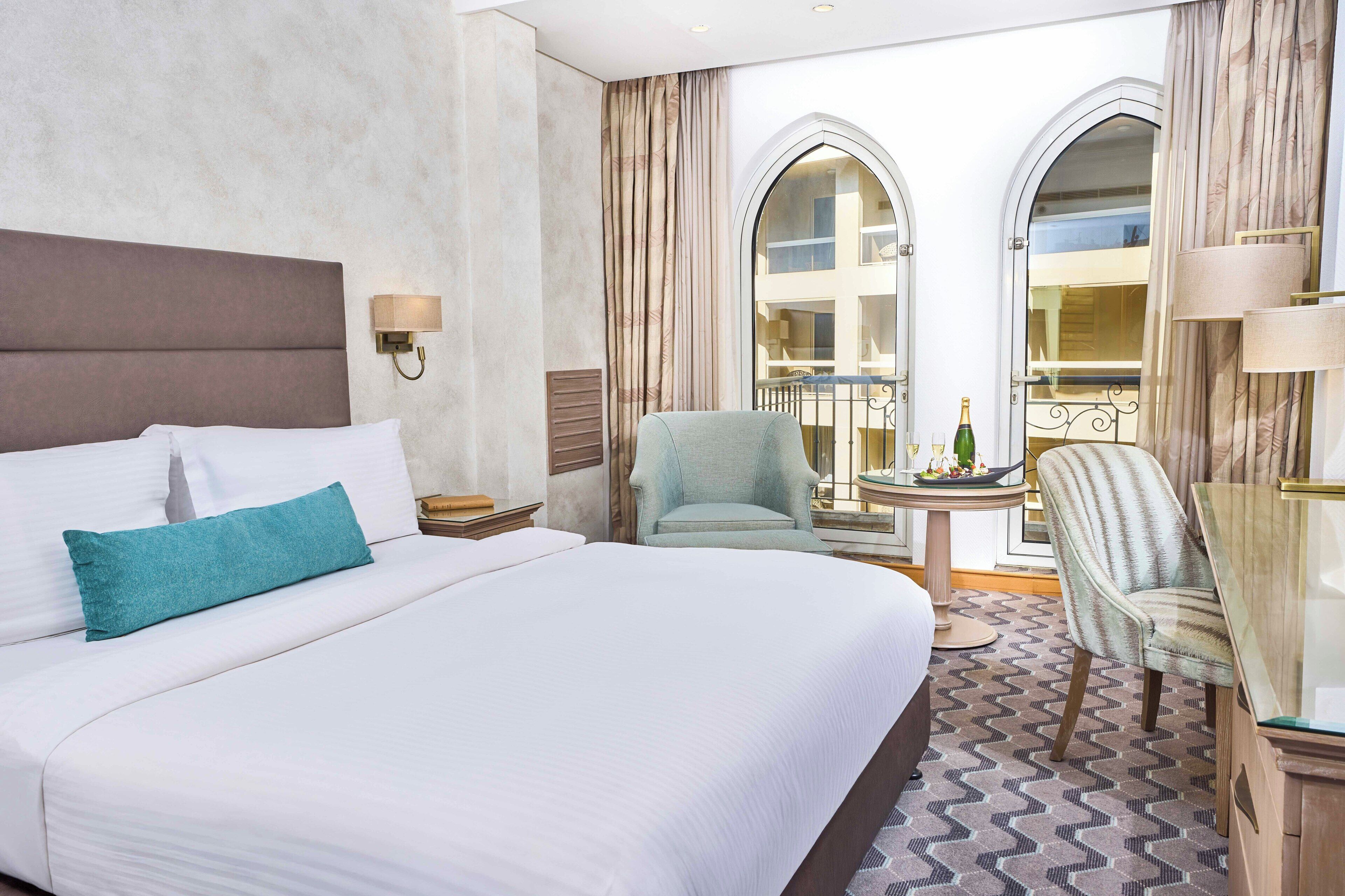 Una habitación de hotel con una cama grande, un área de descanso con una mesa y vistas a la ciudad a través de ventanales arqueados.