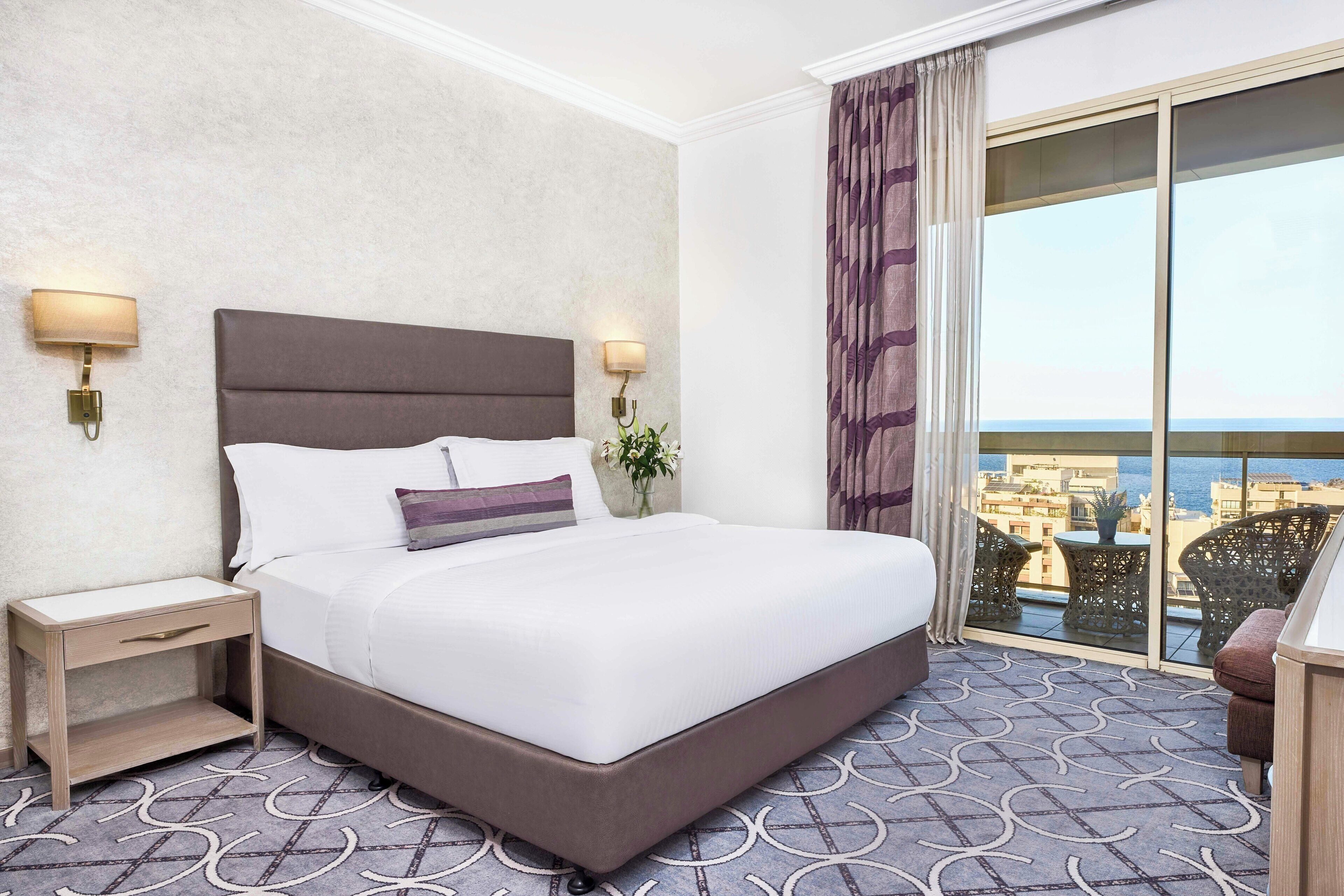 Habitación de hotel con una cama grande, dos mesitas de noche con lámparas, un pequeño atril, una planta en maceta y vistas de la ciudad a través de un amplio ventanal.