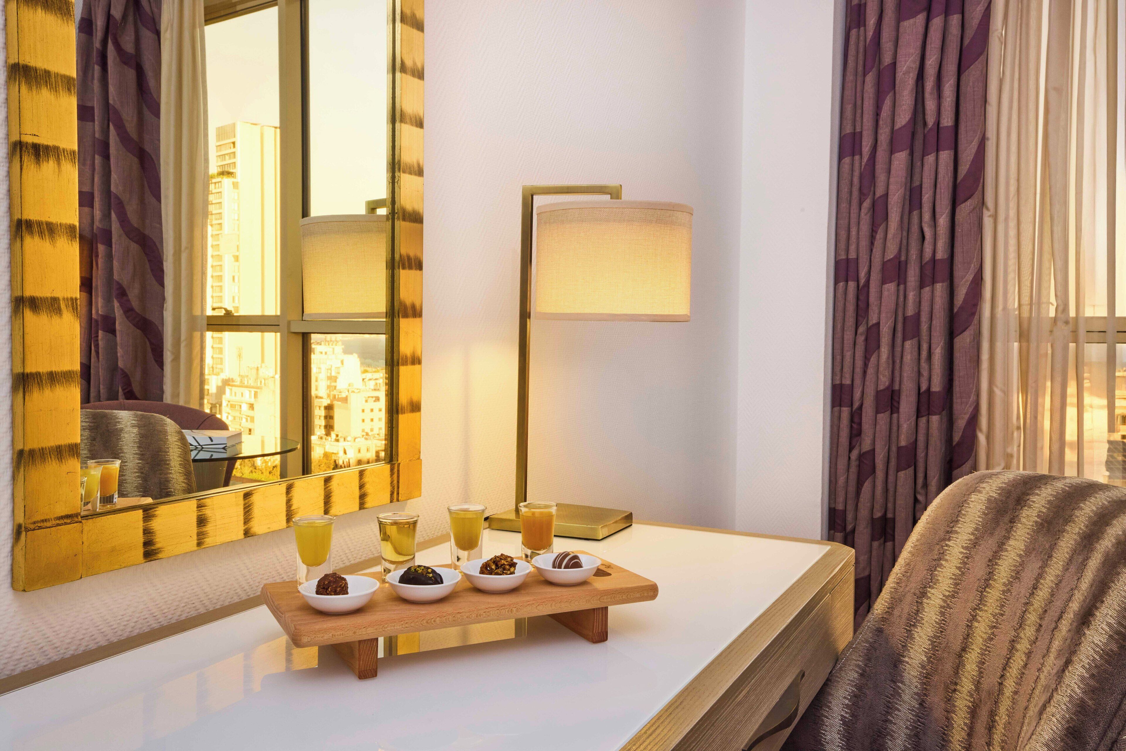A hotel room with a large mirror, a bedside table with a tray of snacks, a lamp, and a view of the city through the window.