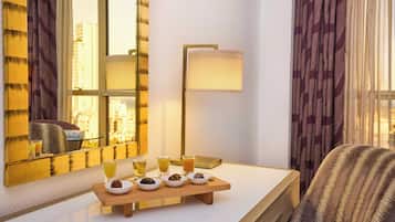 A hotel room with a large mirror, a bedside table with a tray of snacks, a lamp, and a view of the city through the window.