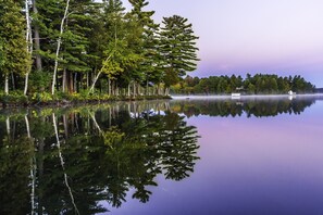 Un lago tranquilo con árboles frondosos reflejándose en el agua, un pequeño muelle y una cabaña a lo lejos.