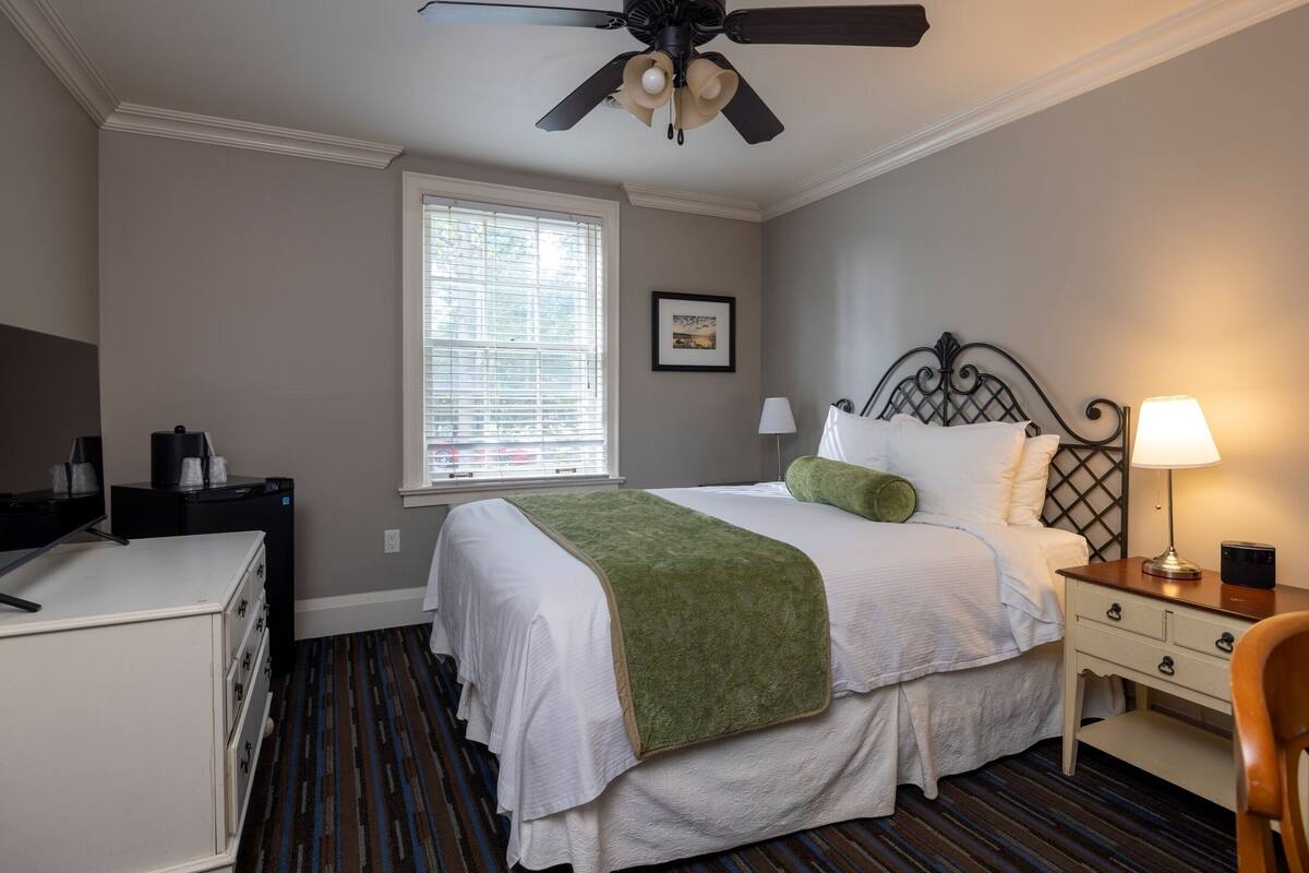 A hotel room with a bed, a desk, a chair, a ceiling fan, a window, and a nightstand with a lamp.