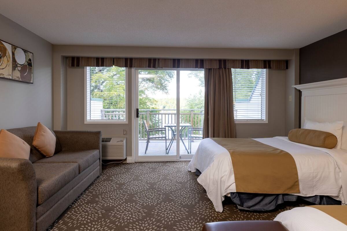A hotel room with a bed, a sofa, a small table, and a balcony with a view of trees.