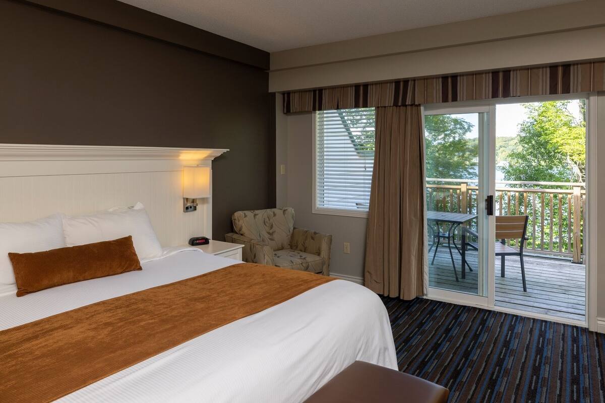 A hotel room with a large bed, a chair, a small table, and a balcony with a table and chairs.