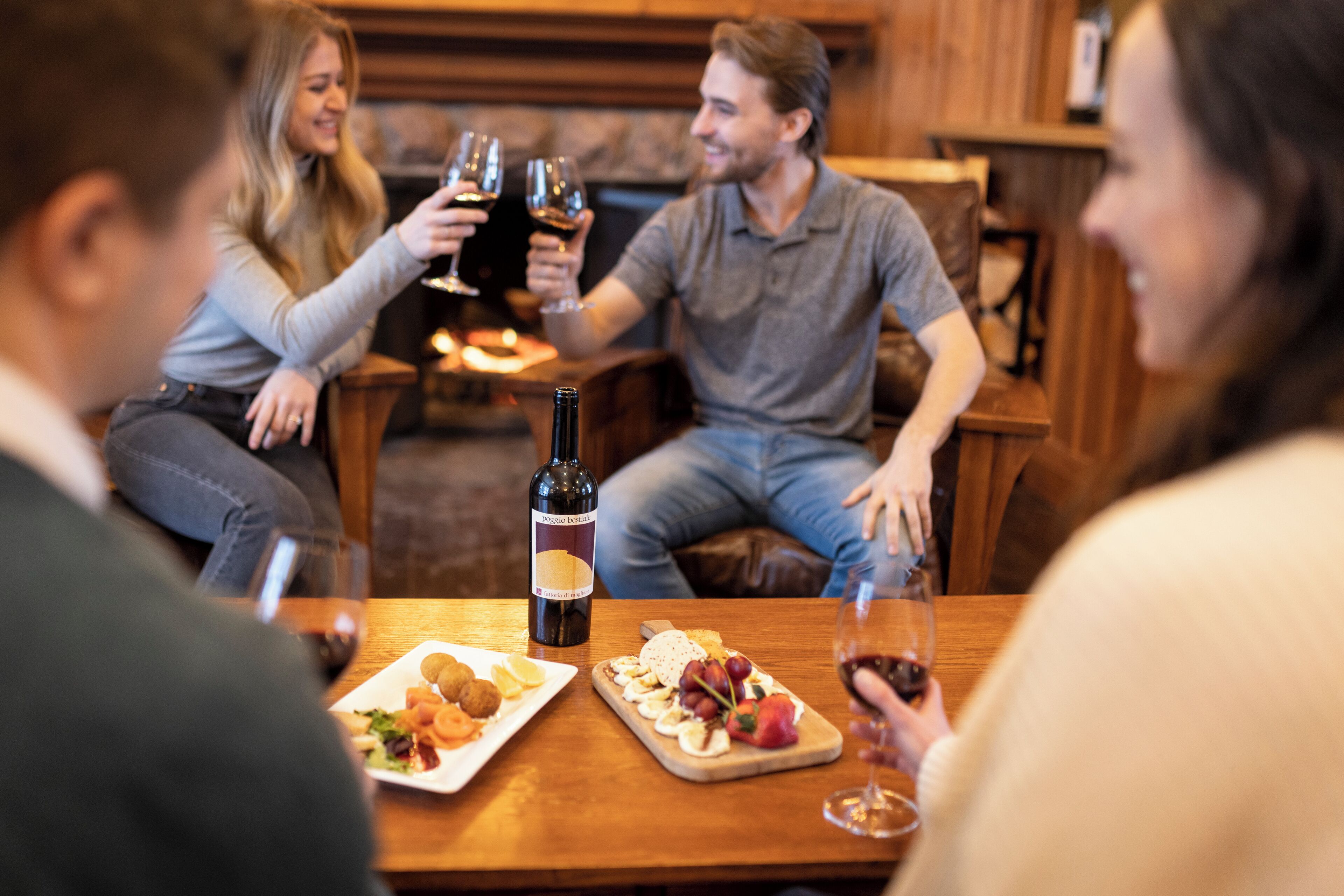 A group of people sitting at a wooden table with wine glasses, a bottle of wine, and plates of food.