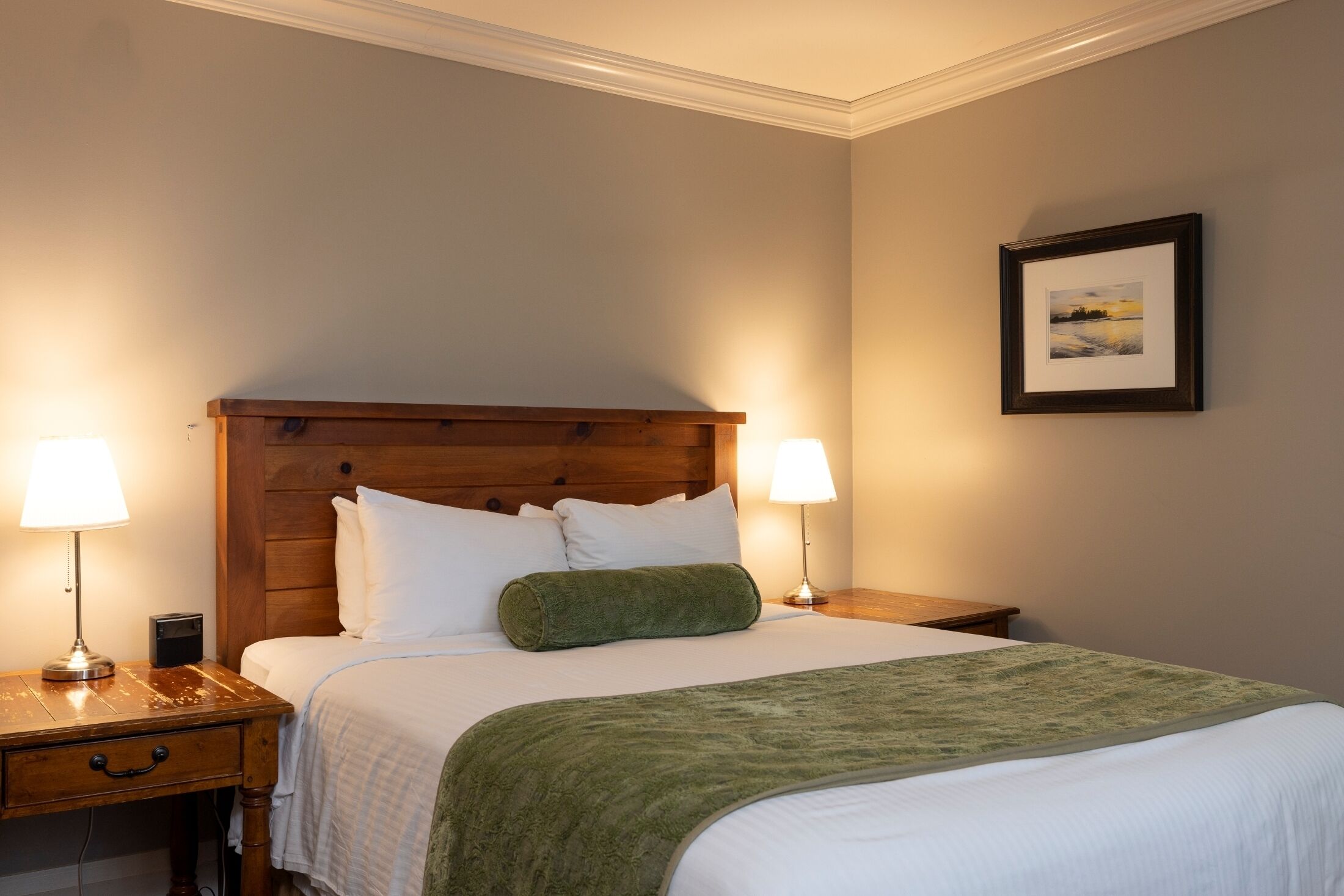 Cama bem arrumada com lençóis brancos e manta verde, com duas mesas de cabeceira com luminárias, num quarto com cabeceira de madeira e quadro na parede.