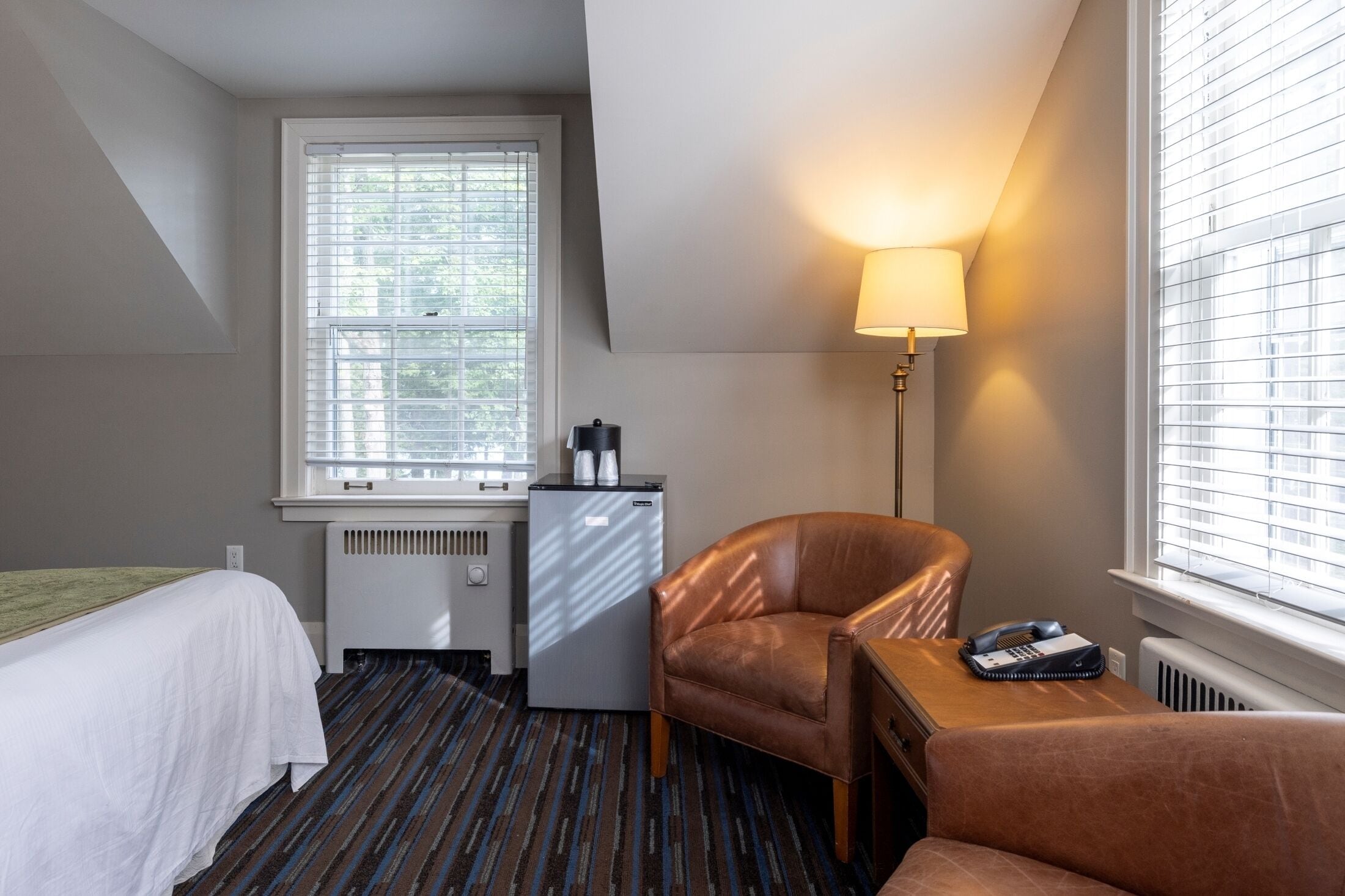 A hotel room with a bed, a desk with a lamp, a chair, and a radiator.