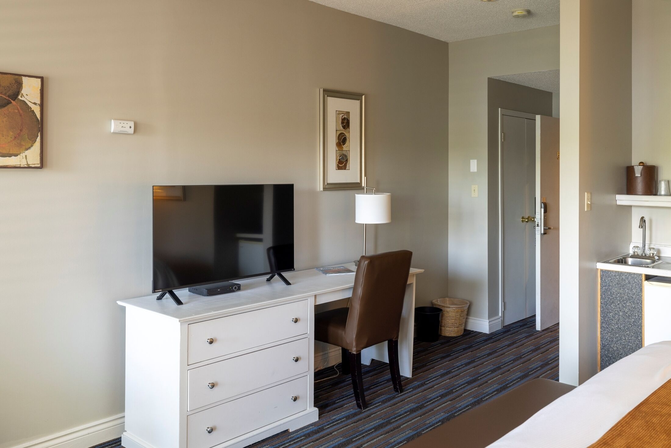 A hotel room with a white desk, a flat-screen TV, a brown leather chair, a framed picture on the wall, and a small bathroom area with a sink and mirror.