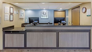 Una recepción con un letrero que dice "Quality by Choice Hotel", dos monitores de computadora, un reloj y documentos enmarcados en la pared.