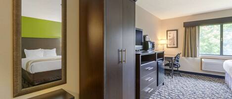 A hotel room with a large wardrobe, a desk with a chair, a bed, a mirror, and a window with curtains.