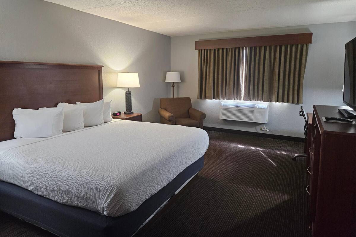 A hotel room with a large bed, two bedside lamps, a chair, a desk with a computer, and a TV.