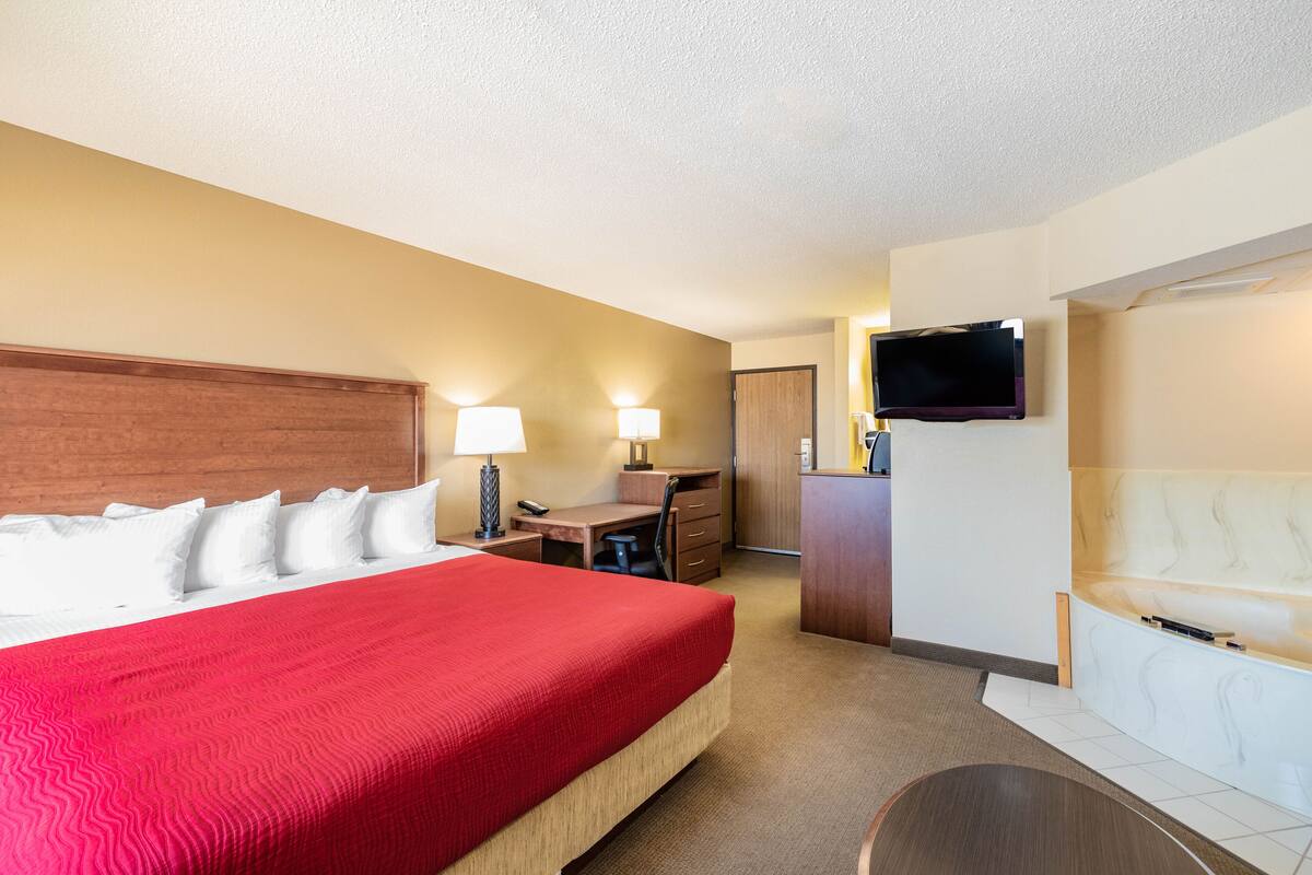 A hotel room with a large bed, a desk, a chair, a TV, and a small seating area.