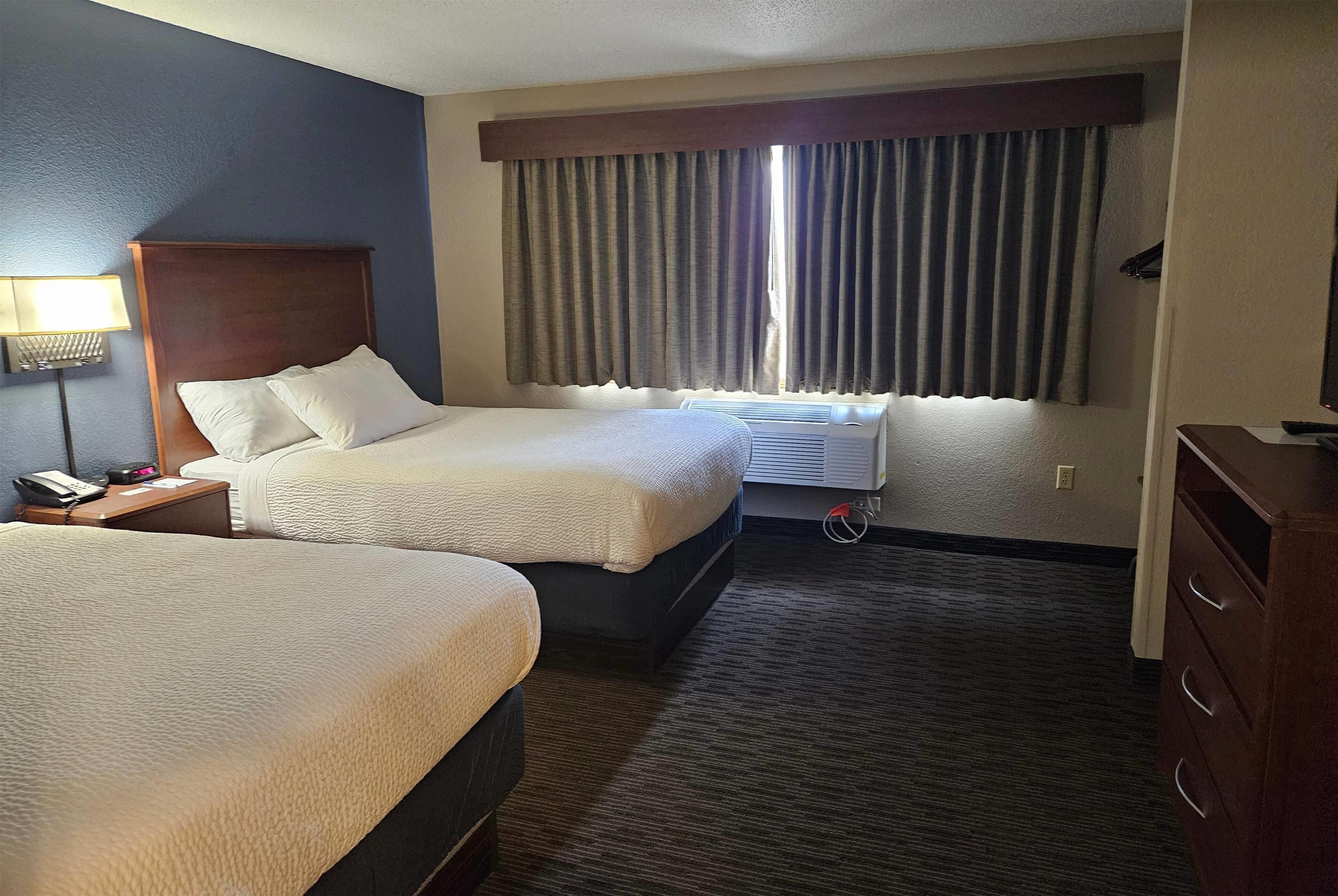 Habitación de hotel con dos camas, una mesita de noche con lámpara, cómoda y unidad de aire acondicionado cerca de la ventana.
