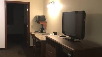 A compact hotel room with a kitchenette, a desk with a computer and a flat-screen TV, and a door leading to another room.