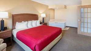 A hotel room with a large bed, a bedside table with a lamp, a nightstand, a bathroom with a bathtub, and a door with frosted glass panels. - AmericInn by Wyndham Marshall (Marshall)