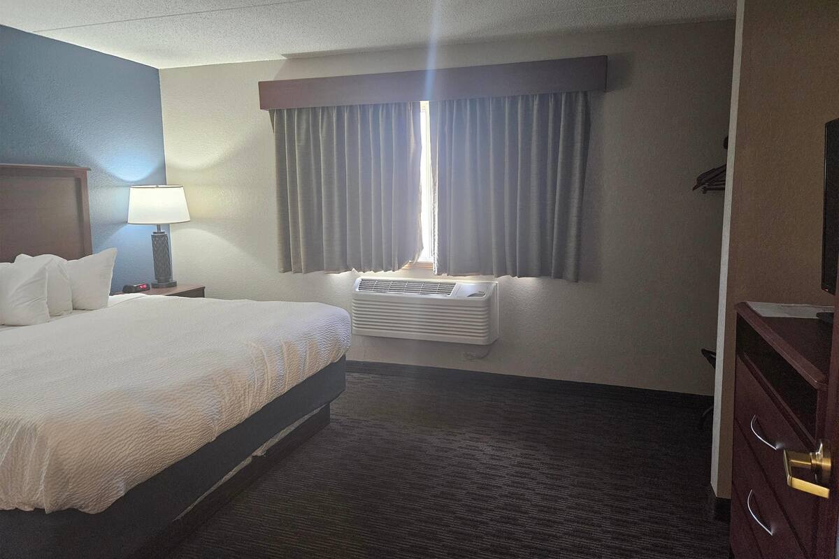 A hotel room with a bed, a nightstand, a lamp, a window with curtains, an air conditioning unit, and a dresser.