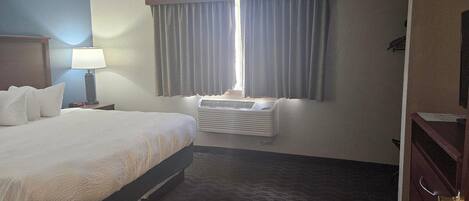 Habitación de hotel con cama, mesita de noche, lámpara, ventana con cortinas, equipo de aire acondicionado y cómoda.