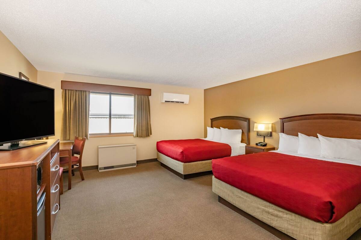 A hotel room with two beds, a TV, a desk, and a window with curtains.