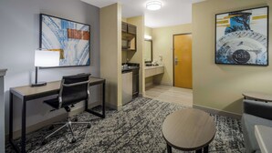 A hotel room with a desk, chair, and a small round table. There is a microwave, a sink, and a mirror in the kitchenette.