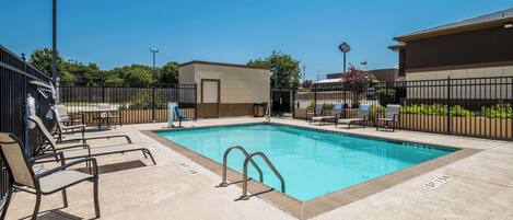 Una piscina rectangular con solárium empedrado, rodeada de sillas de descanso y un pequeño edificio.