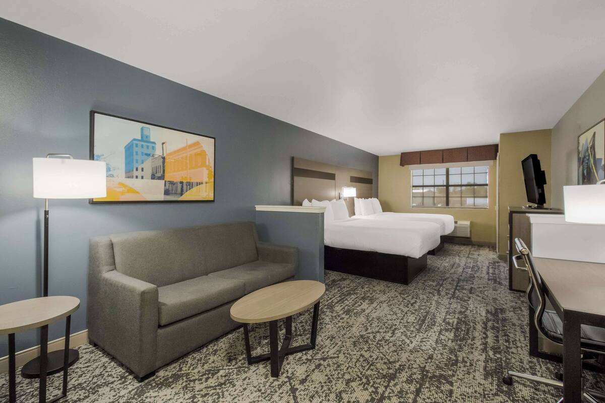 Ein Hotelzimmer mit einer Couch, zwei Einzelbetten, einem Schreibtisch, einem Stuhl, einem kleinen Tisch, einer Lampe und einem Bild an der Wand.