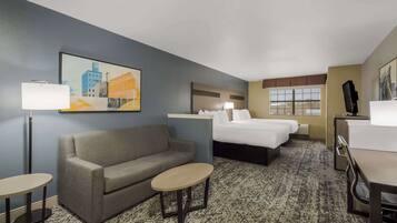 A hotel room with a sofa, two single beds, a desk, a chair, a small table, a lamp, and a painting on the wall.