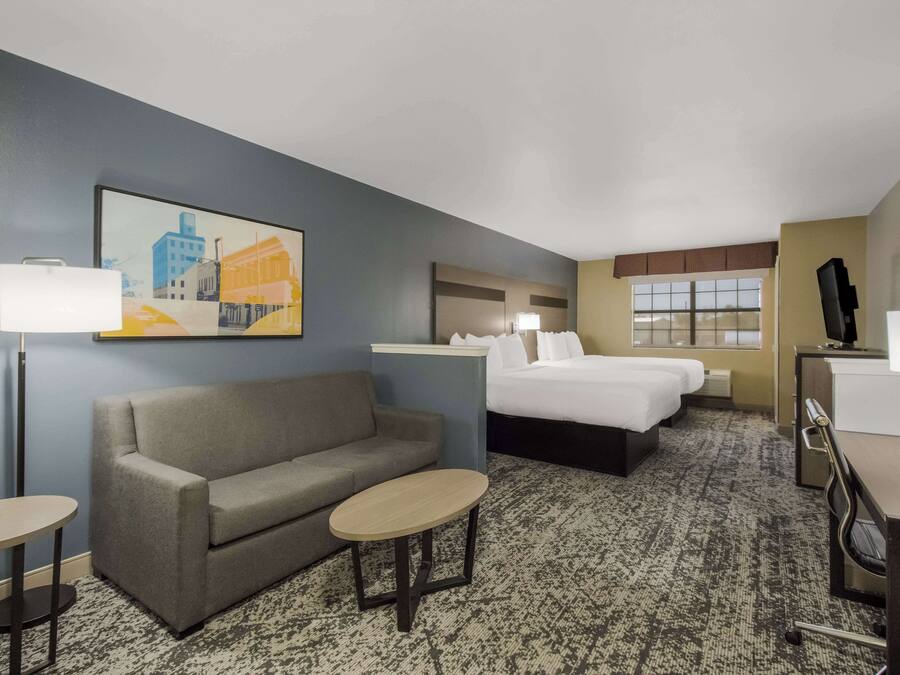 A hotel room with a sofa, two single beds, a desk, a chair, a small table, a lamp, and a painting on the wall.