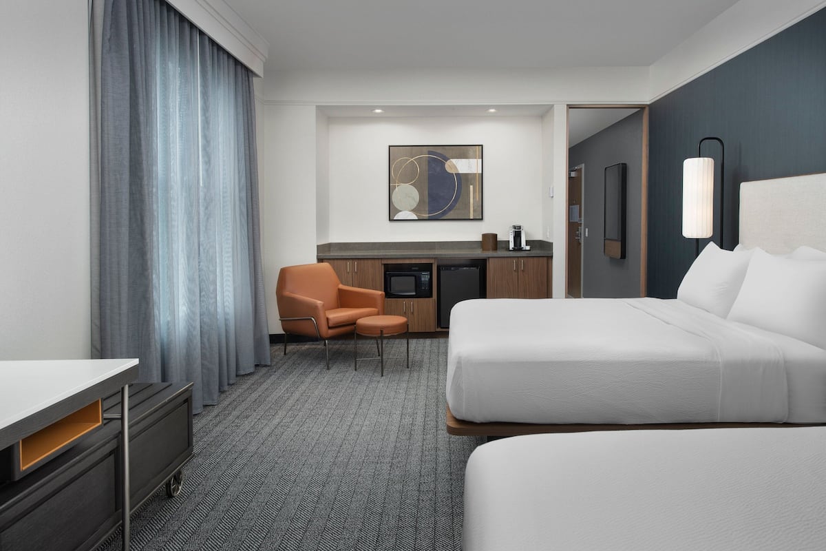 A hotel room with two beds, a desk, a chair, a small kitchenette, and a large window with curtains.
