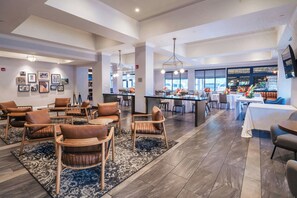 Un amplio vestíbulo de hotel con una zona de estar central, un bar y un comedor con grandes ventanales.