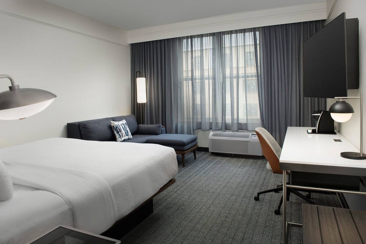 A hotel room with a bed, a desk, a chair, a sofa, a TV, and an air conditioning unit.