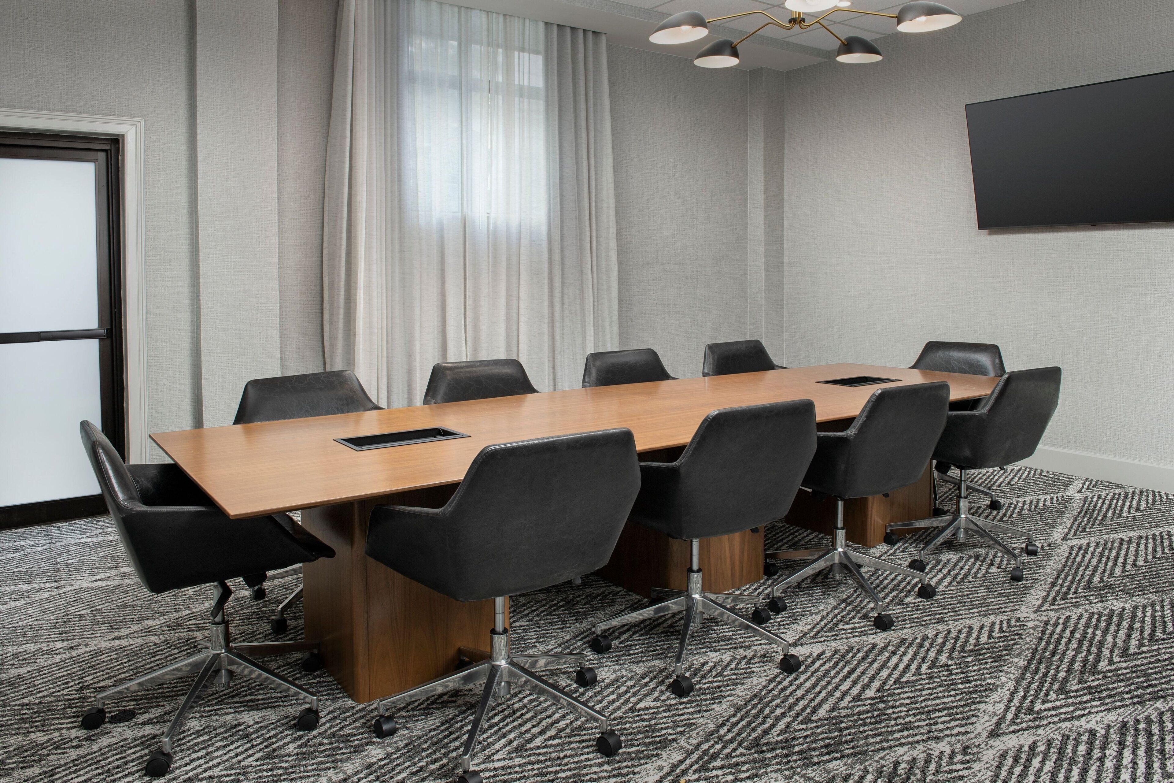 Une salle de conférence avec une longue table, des chaises noires, un sol recouvert de moquette, une grande fenêtre avec des rideaux et un téléviseur à écran plat fixé au mur.