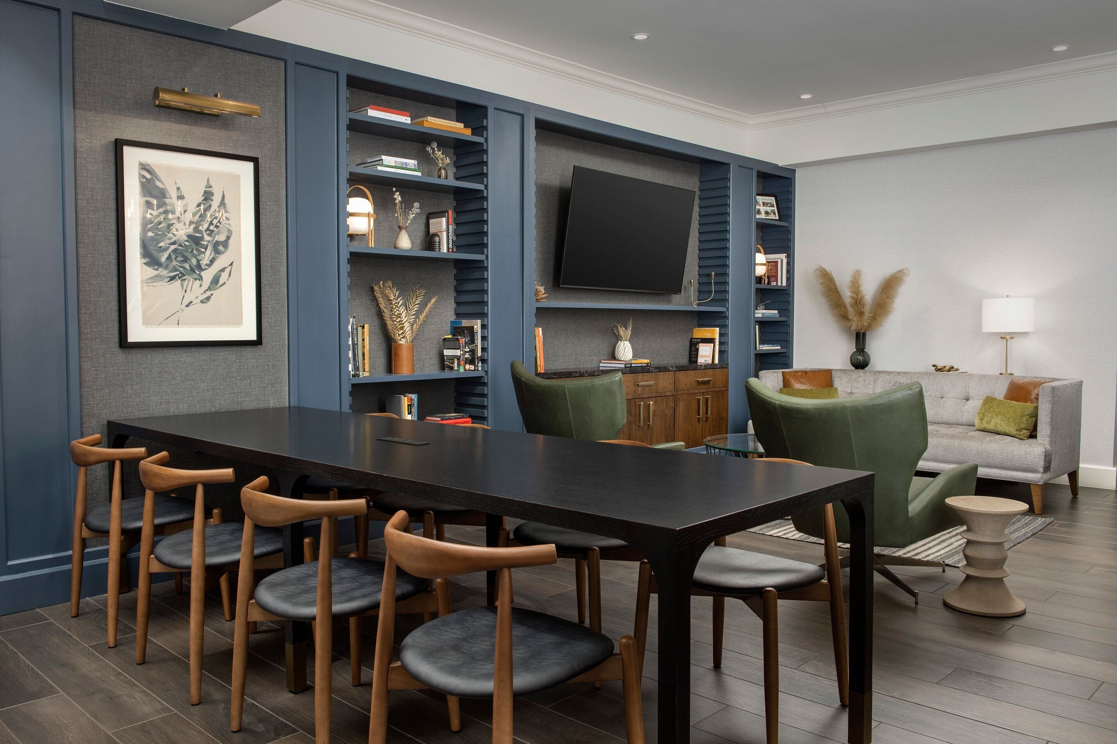 A modern dining area with a dark wooden table and matching chairs, a bookshelf with decorative items, a flat-screen TV mounted on the wall, and a grey sofa with a lamp.