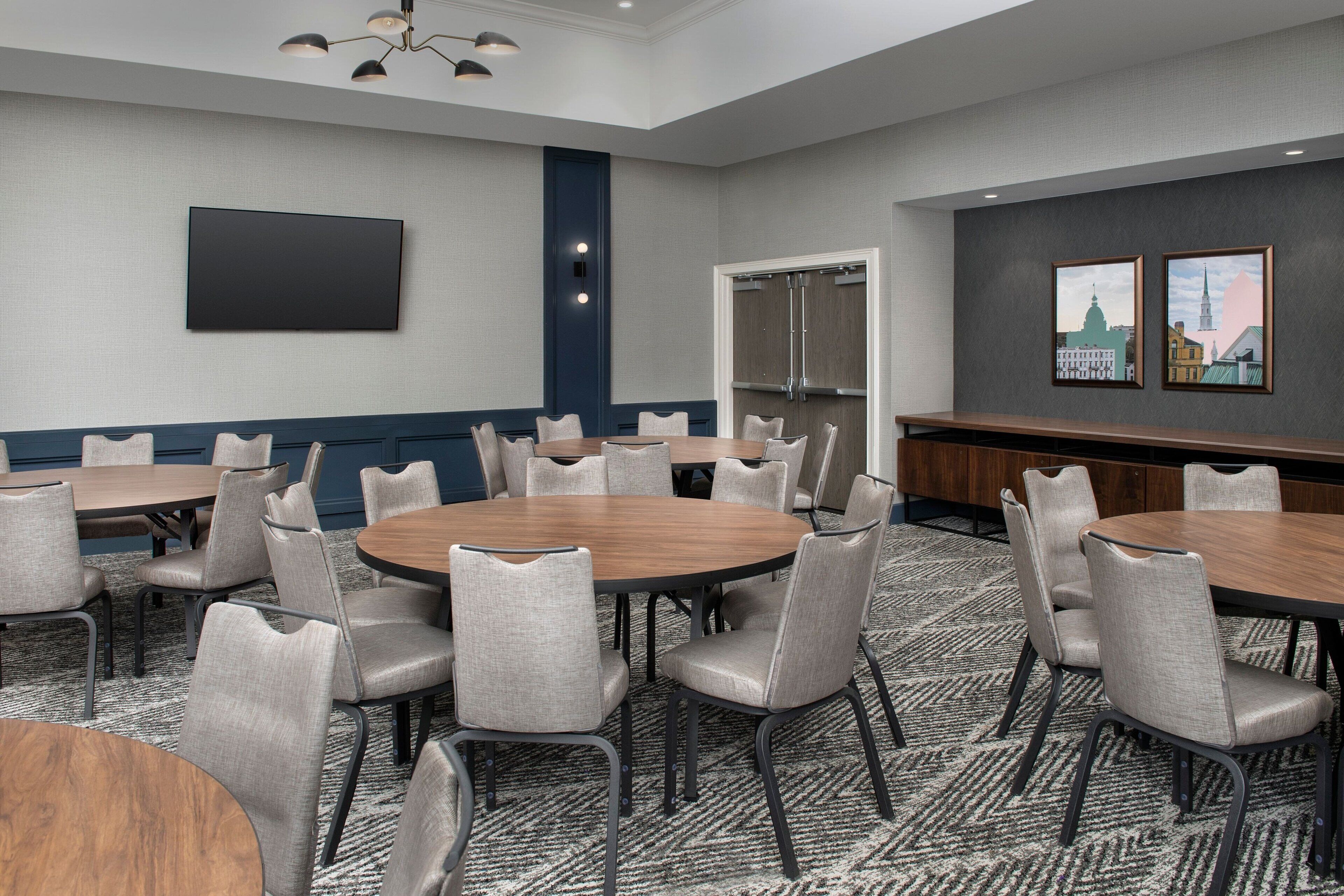 A conference room with round tables and chairs, a flat-screen TV, a door, and two framed pictures on the wall.