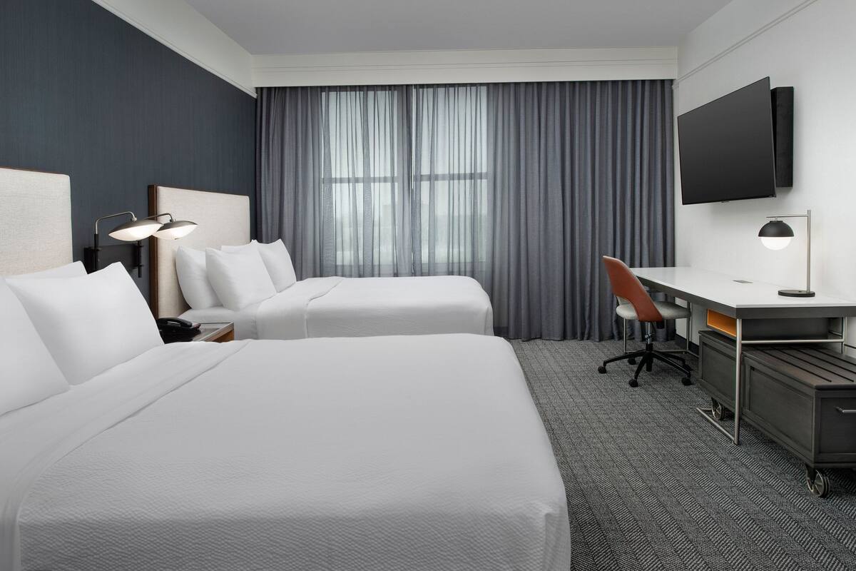 A hotel room with two beds, a desk, a chair, a flat-screen TV, and a large window with curtains.