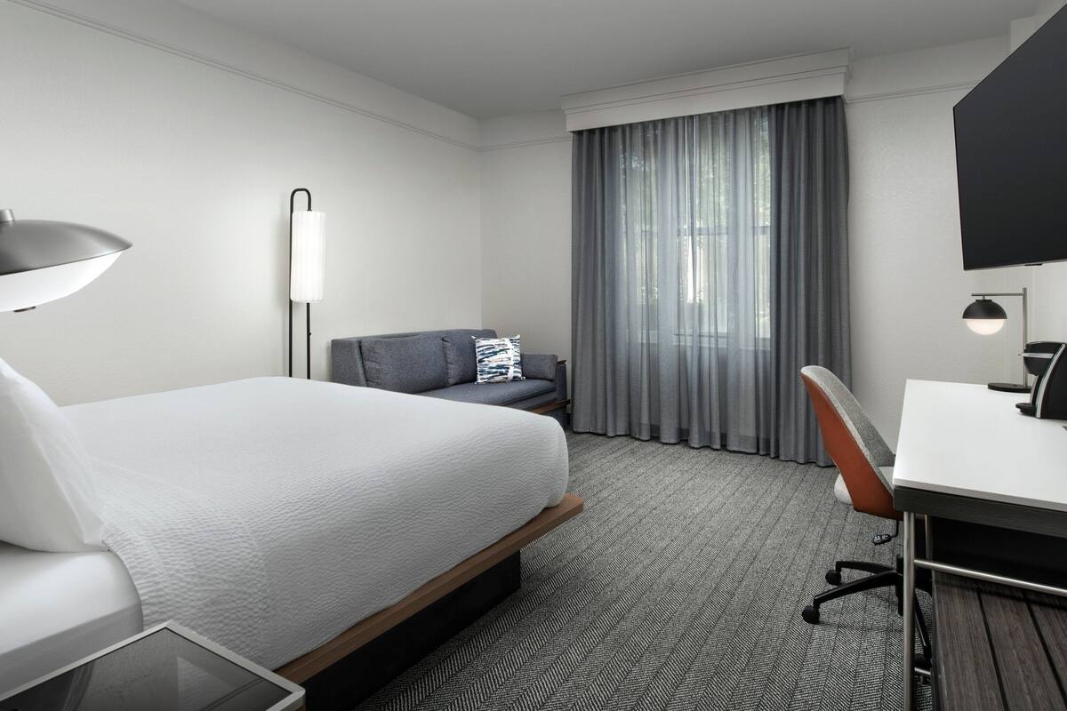 A modern hotel room with a large bed, a desk, a chair, a lamp, and a large window with curtains.