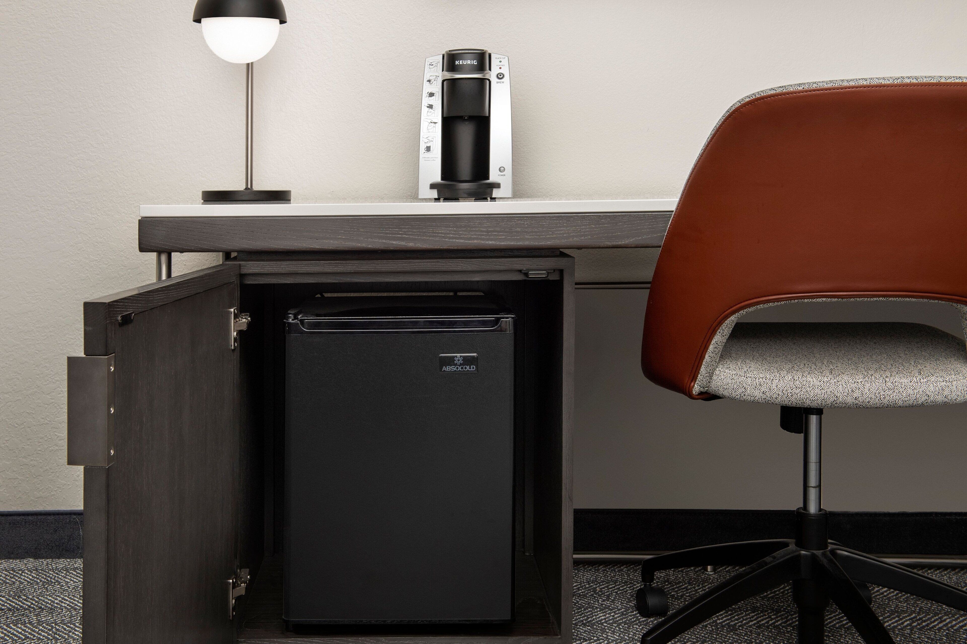 Un mini-bar compact de couleur sombre, équipé d’une machine à café et d’une lampe sur un bureau.