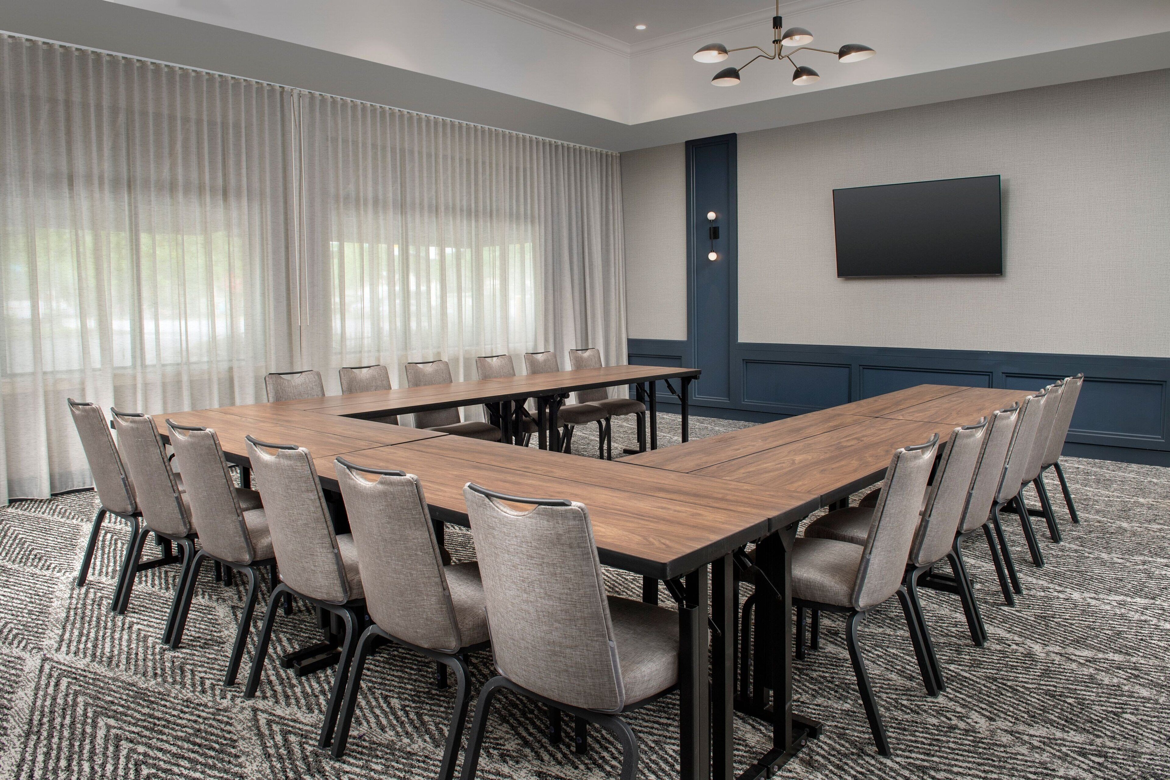 A conference room with a long table and chairs, a large window with curtains, a flat-screen TV, and a patterned carpet.