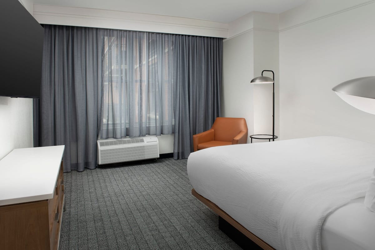 A hotel room with a bed, a desk, an orange chair, a floor lamp, and a window with curtains.