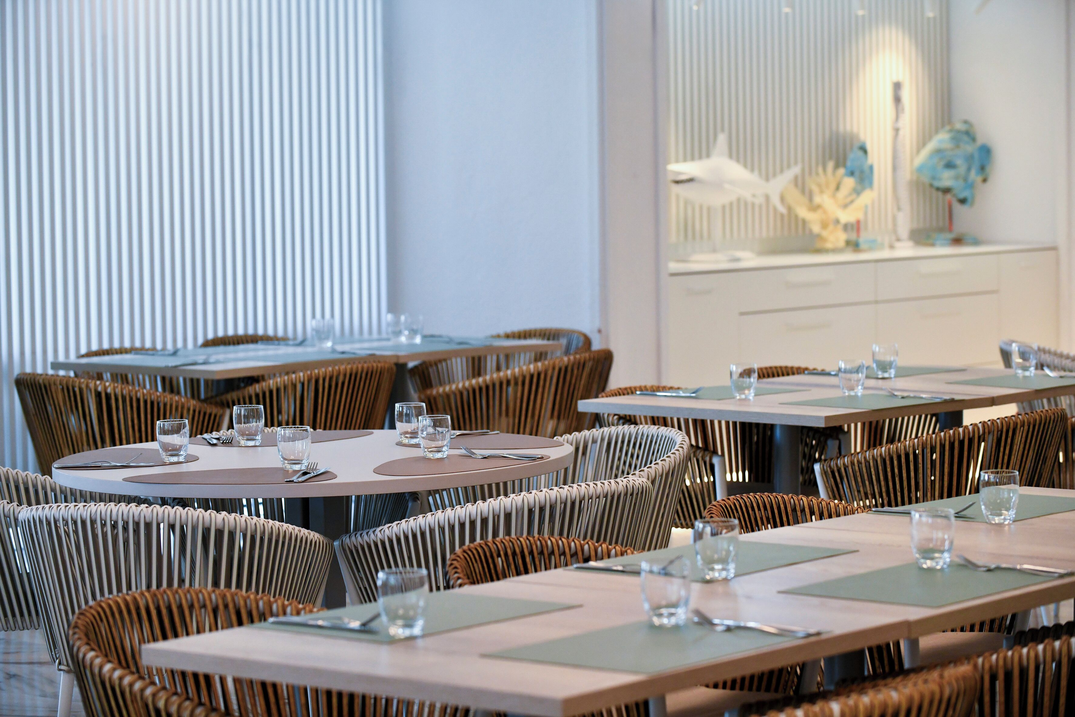 Un restaurante con mesas redondas y sillas de mimbre, con una paleta de colores neutros y una pared decorativa con una escultura.