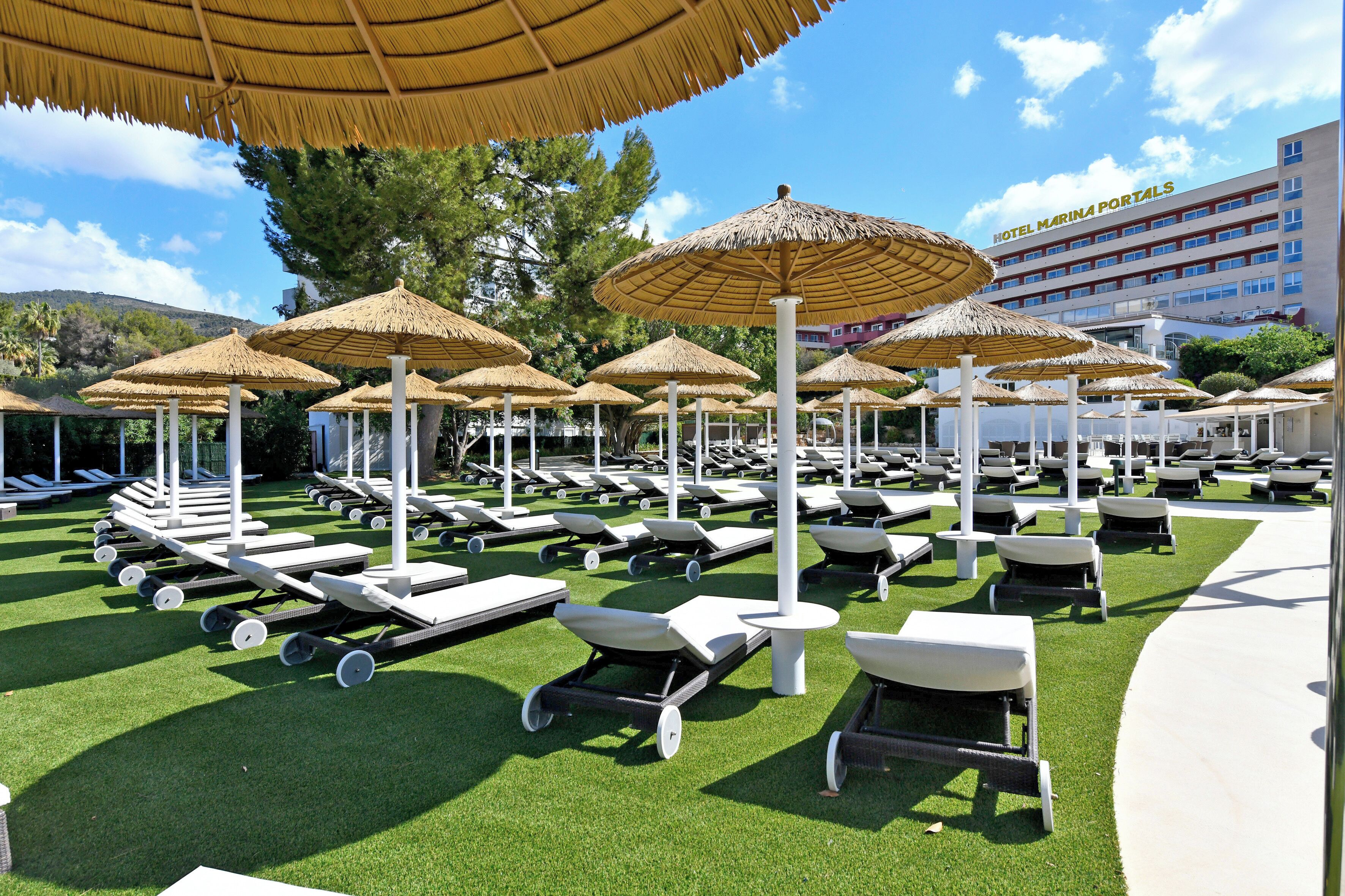 Un espace aménagé au bord de la piscine, avec des chaises longues et des parasols en chaume, un bâtiment portant l’inscription « Hôtel Marina Portals » en arrière-plan, et un ciel dégagé.