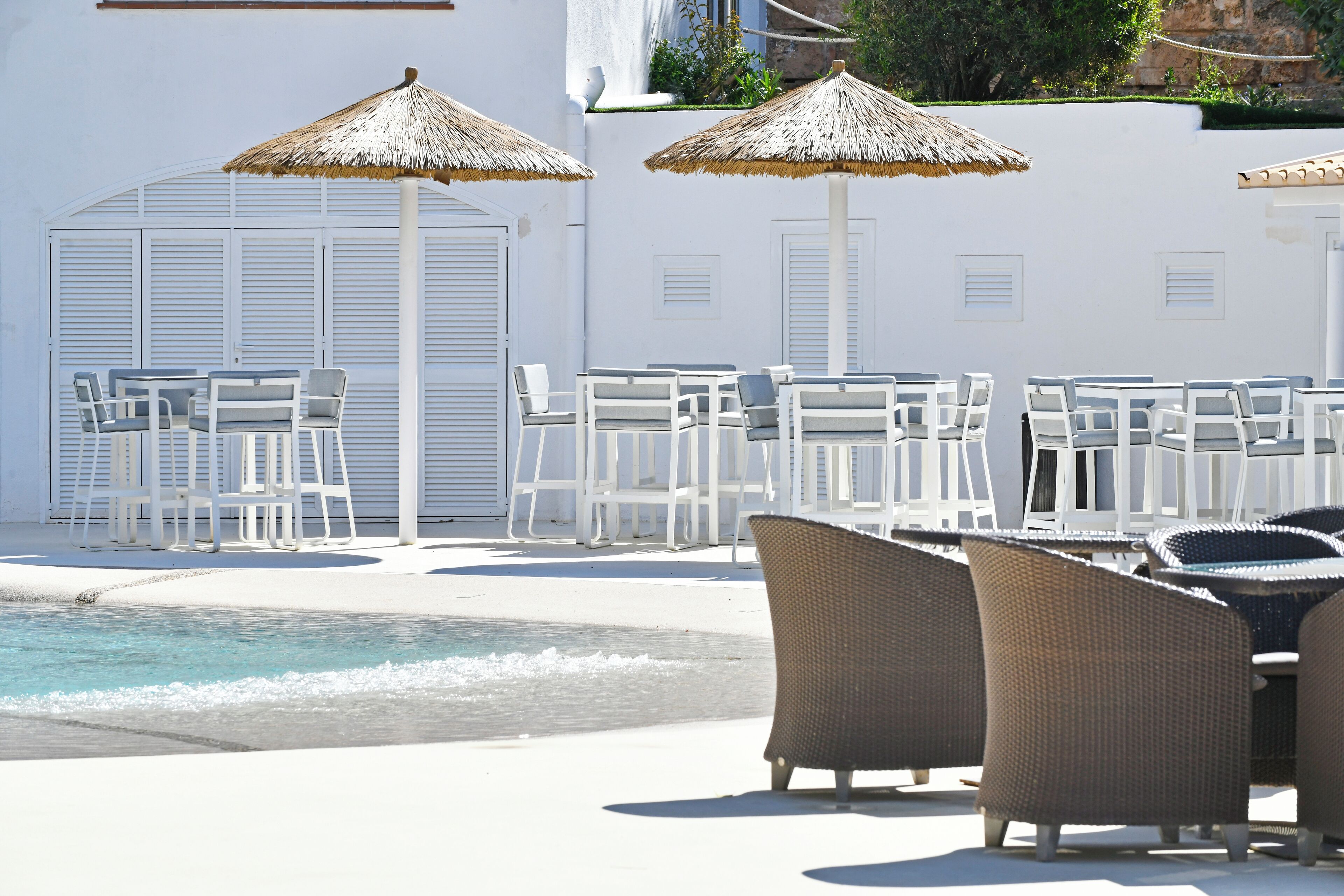 A poolside area with white lounge chairs and thatched umbrellas, a clear pool, and a white building with shutters.